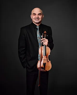 Adrian Petrutiu é um homem branco, calvo. Ele usa camisa e paletó pretos, uma gravata azul e dourada, e segura seu violino.