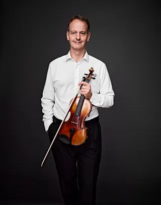 Andreas Uhlemann é um homem branco, de cabelos grisalhos. Ele veste uma camisa branca, sorri e segura um violino.