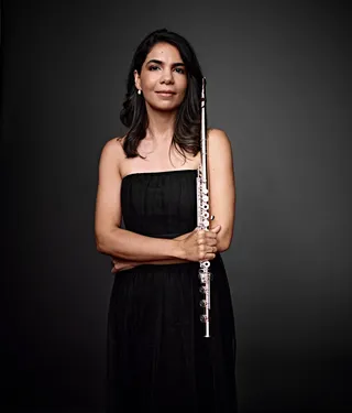 Claudia Nascimento é uma mulher branca, com cabelos e olhos castanhos. Ela usa um vestido preto e está segurando uma flauta.  