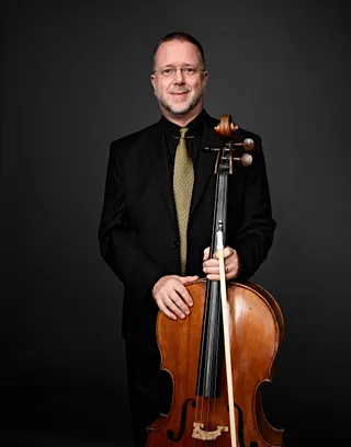 Douglas Kier é um homem branco, de cabelos curtos e barba branca. Ele usa uma gravata amarela, camisa e paletó pretos, e segura um violoncelo.