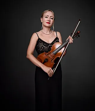 Elena Klementieva é uma mulher branca, de cabelos presos e loiros e batom vermelho. Ela usa um vestido de alças, um colar de pérolas e segura o arco de um violino.