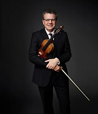Emmanuele Baldini é um homem branco, de óculos e cabelos grisalhos. Ele veste terno, gravata vermelha e camisa branca, e segura um violino.