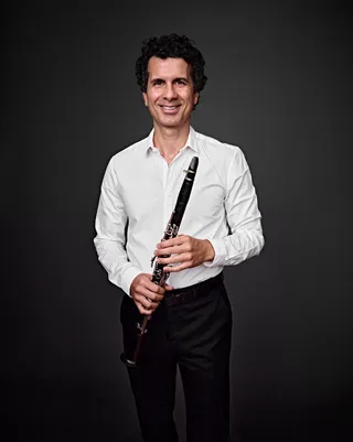 Giuliano Rosas é um homem branco, de cabelos encaracolados, ele está sorrindo e segura um clarinete.. 