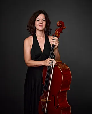 Heloisa Meirelles é uma mulher branca, de cabelos castanhos na altura do ombro. Ela veste um vestido preto decotado e segura um violoncelo.
