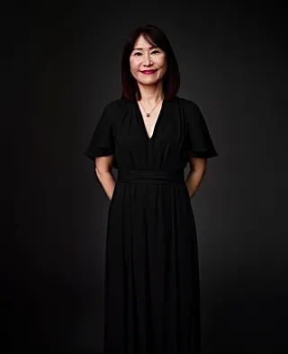 Ji Sook Chang é uma mulher coreana, de cabelos na altura dos ombros e franja. Ela usa batom rosa, vestido preto e um colar dourado com um pingente.