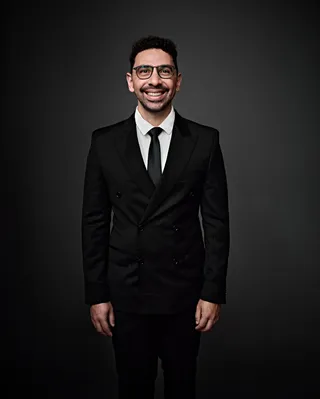 João Vitor é um homem branco, na faixa dos 30 anos, com cabelos curtos pretos. Ele veste camisa branca, e gravata e terno pretos. João usa óculos e sorri para a foto.