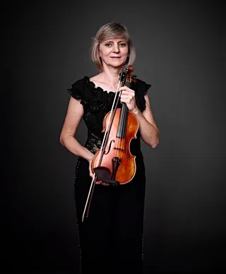 Katia Spassova é uma mulher branca, de cabelos curtos e loiros. Ela veste um vestido preto com babados e segura um violino.