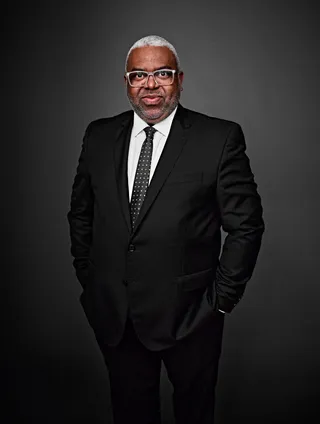 Moisés Téssalo é um homem negro, com cabelos curtos grisalhos. Ele veste camisa branca, gravata e paletó pretos, e usa óculos.