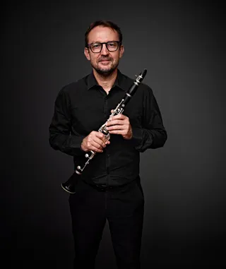 Ovanir Buosi é um homem branco, de barba e cabelos castanhos. Ele usa óculos, usa camisa preta e segura um clarinete.