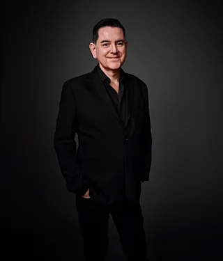 Foto de Paulo Cerqueira, homem branco, de cabelos curtos e pretos. Veste camisa preta, que está com o último botão do colarinho aberto. Ele sorri para a foto. 