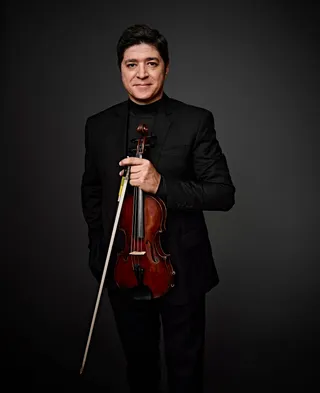 Paulo Paschoal é um homem branco, de cabelos castanhos. Ele veste camisa e paletó pretos, e segura um violino.