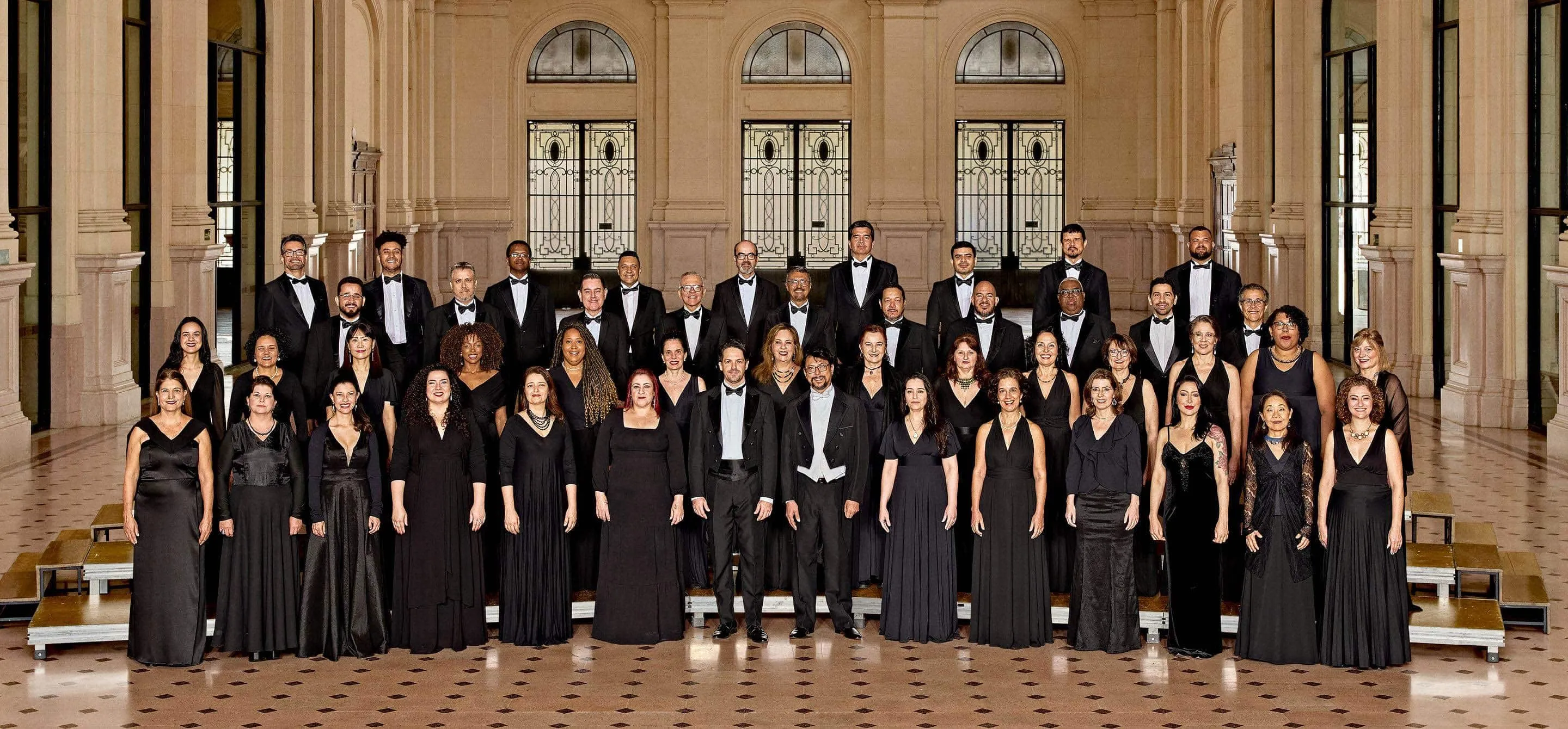 Coro da Osesp, composto por 45 homens e mulheres. Na imagem, eles estão no palco da Sala São Paulo, dispostos em 4 fileiras, e vestidos com trajes de concerto.