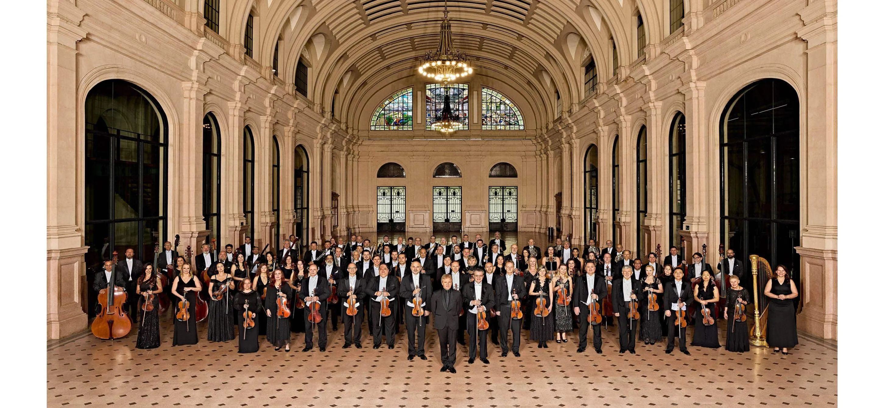 Os músicos da Orquestra estão posicionados na Estação das Artes da Sala São Paulo, um grande salão com colunas beges e chão com desenhos com de rosa. Os músicos vestem preto e estão segurando seus instrumentos.