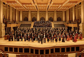 Os músicos da Orquestra e do Coro estão na sala de concertos da Sala São Paulo. Eles vestem preto, seguram seus instrumentos e sorriem.