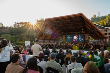 Pessoas aplaudem uma orquestra em um palco ao ar livre, com o sol se pondo e árvores ao fundo.