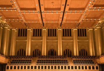 A imagem mostra a visão da Sala São Paulo vista do palco, com o teto móvel, as colunas iluminadas e as cadeiras de madeira amarela e estofado azul. 