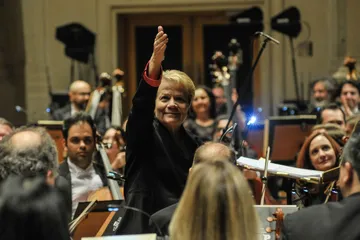 Marin Alsop é uma mulher branca e loira, ela veste blazer preto com os punhos vermelhos.