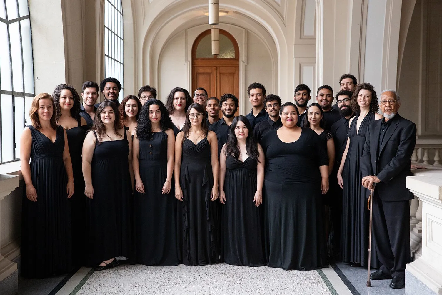 Os coristas da Academia de Música estão posicionados em uma escada no Salão São Paulo. Todos estão vestindo roupas pretas e sorrindo.