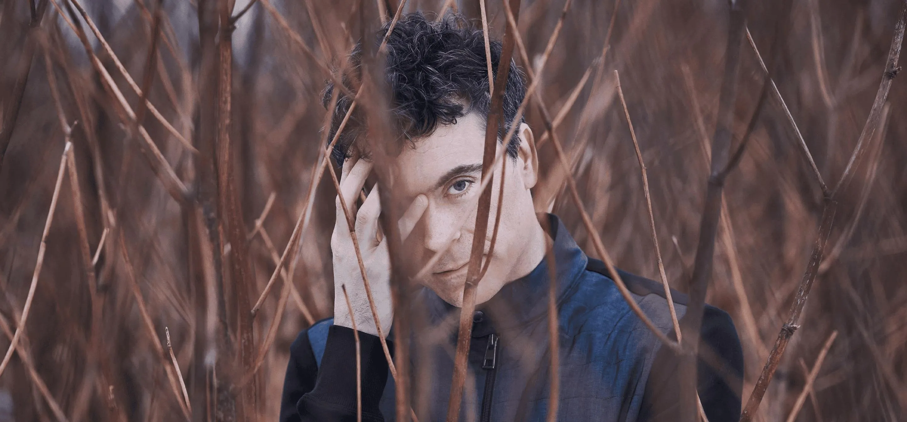 Paul Lewis é um homem branco de olhos azuis e cabelo castanho. Ele veste azul e está com o rosto encoberto por galhos