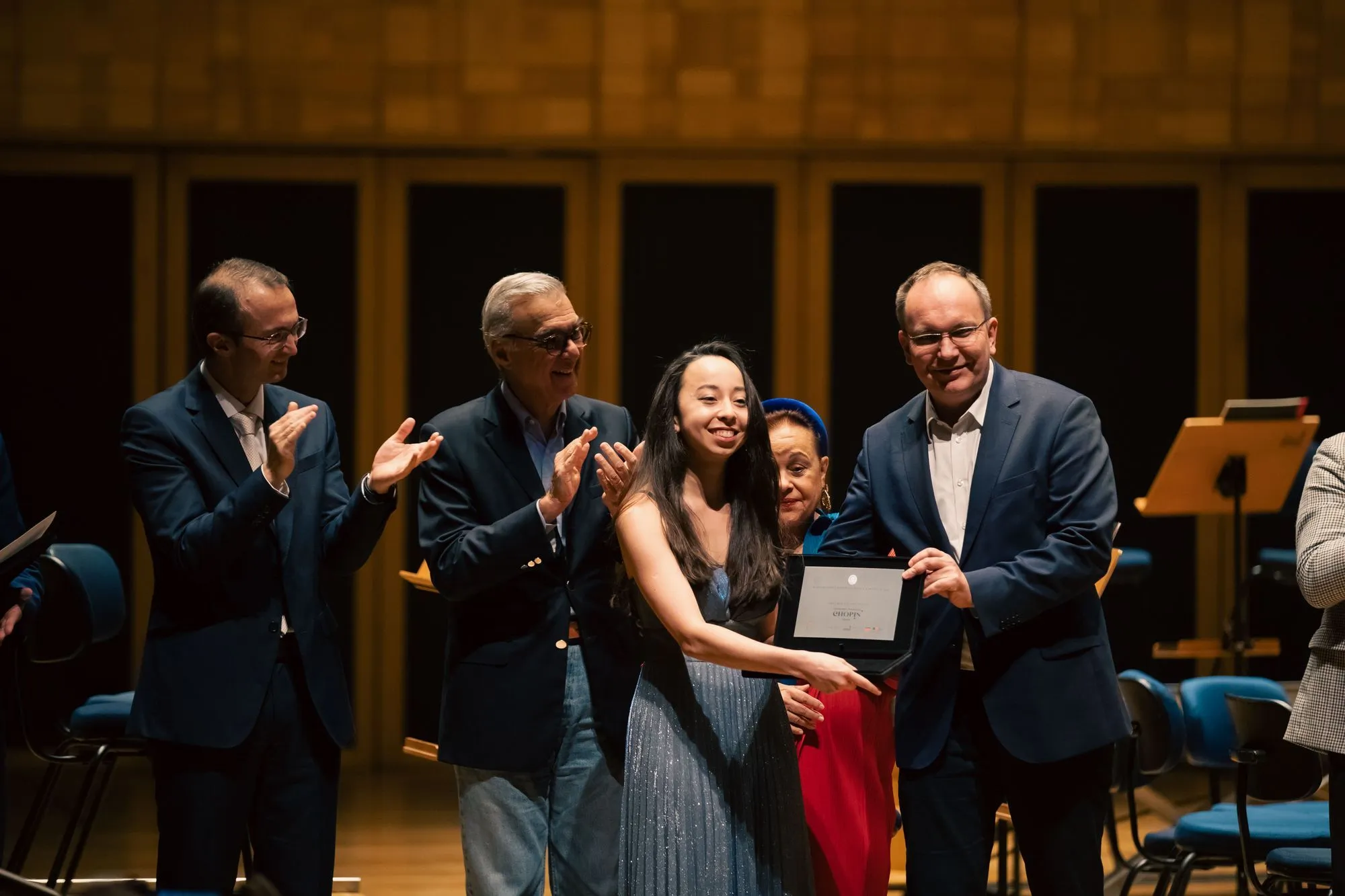 De pé, no palco da Sala São Paulo, Ingrid Uemura. Ela é uma mulher jovem, amarela, de pele clara, cabelos longos e pretos. Usa um vestido longo azul claro, com brilhos e sem mangas.