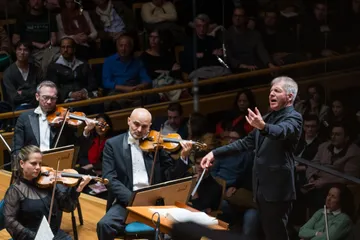 Thierry Fischer é um homem branco, de cabelos grisalhos. Ele rege a Osesp e está com uma expressão emocionada.