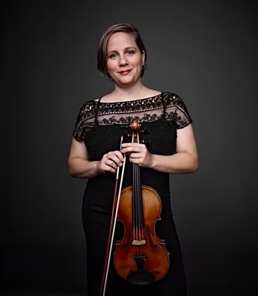 Amanda Martins é uma mulher branca, de cabelos loiros curtos e olhos azuis. Ela está usando uma blusa de renda preta e segurando um violino.