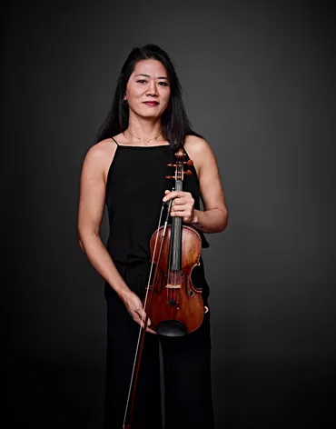 Camila Yasuda é uma mulher asiática, de cabelos longos e pretos. Ela está usando um vestido preto e segurando um violino.