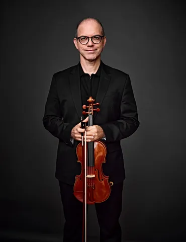 Cesar A. Miranda é um homem branco, calvo, com óculos de armação preta. Ele está usando uma camisa preta e um paletó, e segurando um violino.