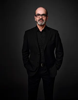 Fábio Vianna é um homem branco, calvo e com barba. Ele está usando óculos e uma camisa preta, com a mão apoiada no queixo.