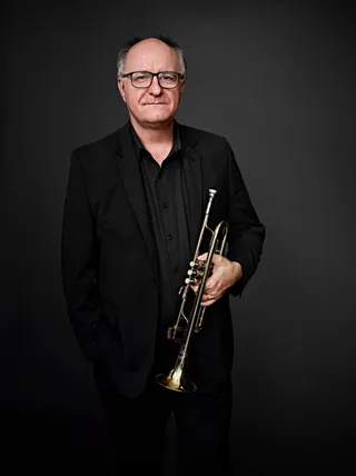 Fernando Dissenha is a white man, bald, and wearing glasses. He is dressed in a black shirt and jacket and is holding a trumpet.