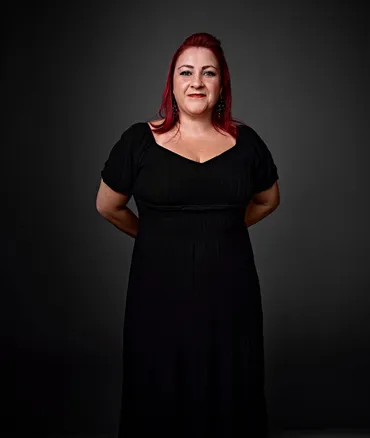 Flavia é uma mulher branca, de olhos verdes e cabelos ruivos. Ela tem um piercing na sobrancelha direita. Está usando um vestido preto e brincos grandes nas cores preta e prata.