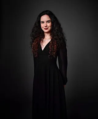 Giulia Moura é uma mulher branca jovem, com cabelo preto, longo e cacheado. Ela está olhando diretamente para frente, sorrindo, e usando um vestido preto e um colar prateado.