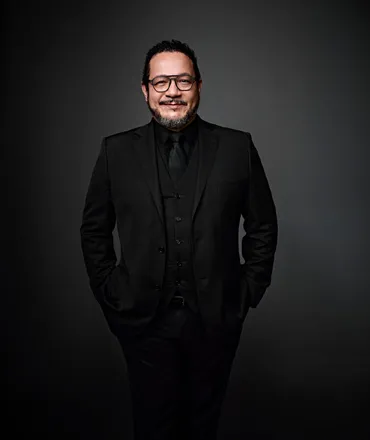 Israel Mascarenhas é um homem com cabelo preto, curto e cacheado, barba e bigode. Ele está usando uma camisa preta, gravata e terno, e também usa óculos.