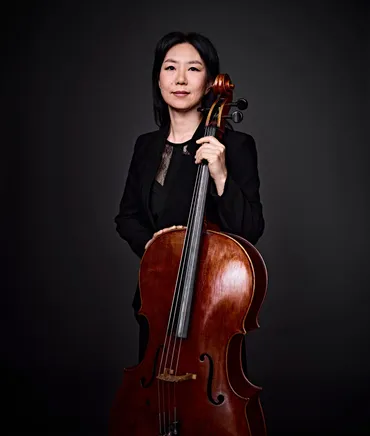 Jin Joo Doh é uma mulher coreana com cabelo preto na altura dos ombros. Ela está vestindo um blazer preto e segurando um violoncelo.