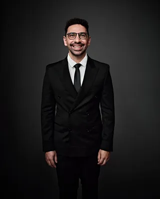 João Vitor é um homem branco, na casa dos 30 anos, com cabelo preto e curto. Ele está usando uma camisa branca, com uma gravata preta e um terno. João está de óculos e sorrindo para a foto.