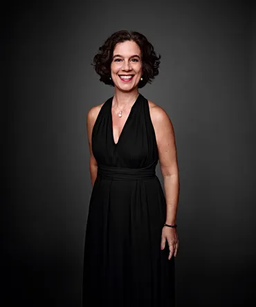 Mariana Valença é uma mulher branca, com cabelo preto, curto e cacheado. Ela está usando um vestido preto e um conjunto combinando de colar e brincos com pérolas.
