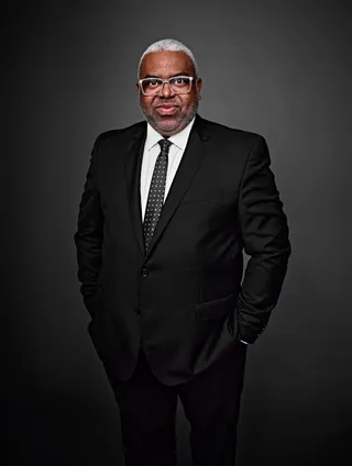 Moisés Téssalo é um homem negro, com cabelo grisalho e curto. Ele está usando uma camisa branca, com uma gravata preta e um blazer, e está de óculos.