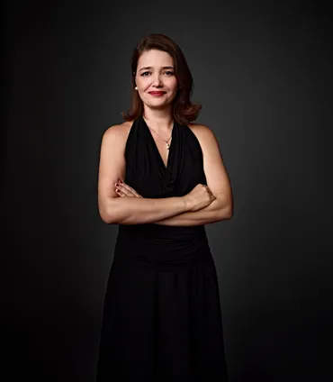 Regiane é uma mulher branca, com olhos claros e cabelo castanho de comprimento médio. Ela está usando um vestido preto, brincos de pérola e um colar de prata com um pingente de pérola. Ela está sorrindo na foto.