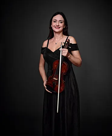 Tatiana Vinogradova é uma mulher branca, com cabelos castanhos longos e olhos claros. Ela está usando um vestido preto decotado e segurando um violino.