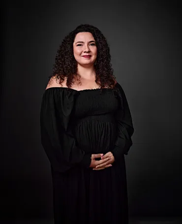 Valquíria é uma mulher branca, com cabelo longo e cacheado de tom escuro. Ela está usando batom rosa e um vestido preto, e está sorrindo na foto.