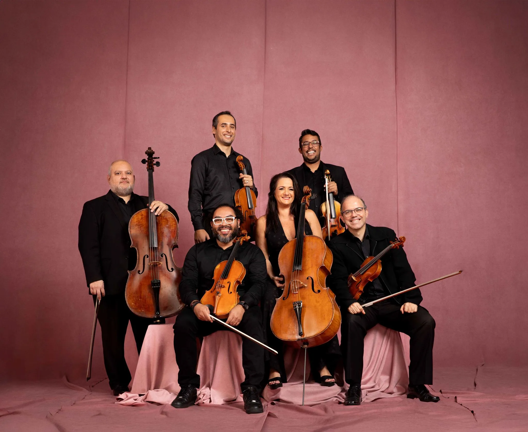 Os músicos estão segurando seus instrumentos em frente à um fundo rosa.