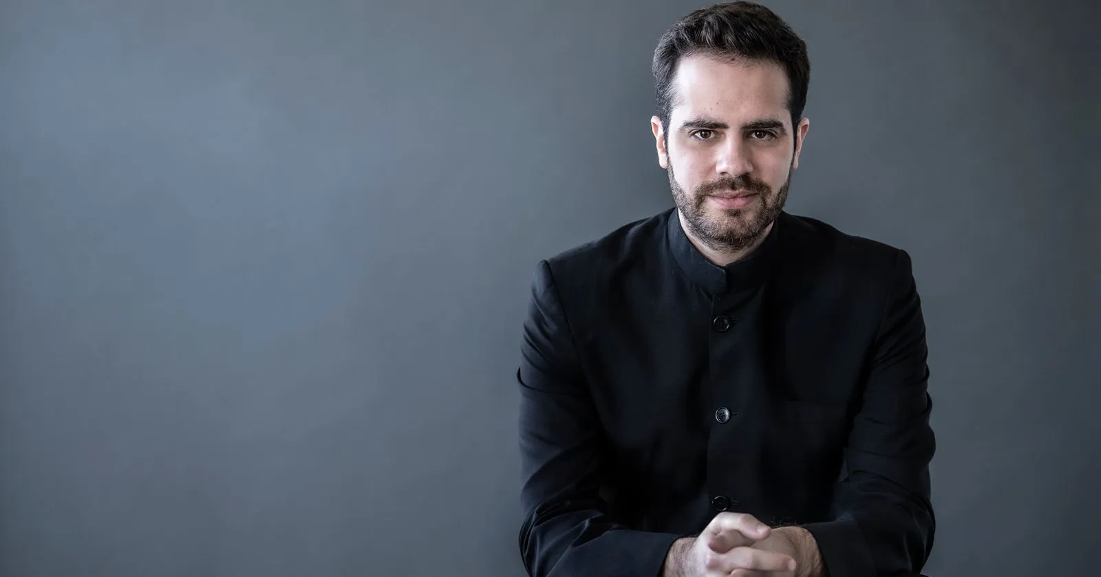 Dinis Souza é um homem branco, de cabelos e barba castanhos, vestindo preto.