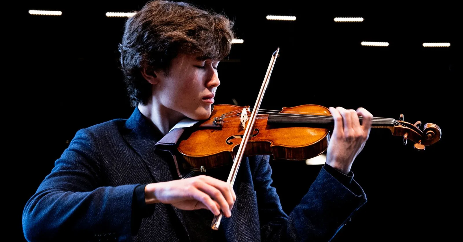 Daniel Lozakovich é um homem branco, de cabelos pretos. Ele está tocando violino.