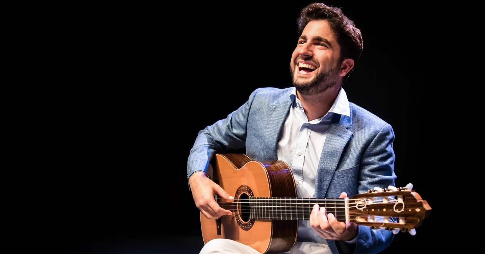 Rafael Aguirre é um homem branco, de cabelos castanhos. Ele toca violão e sorri.