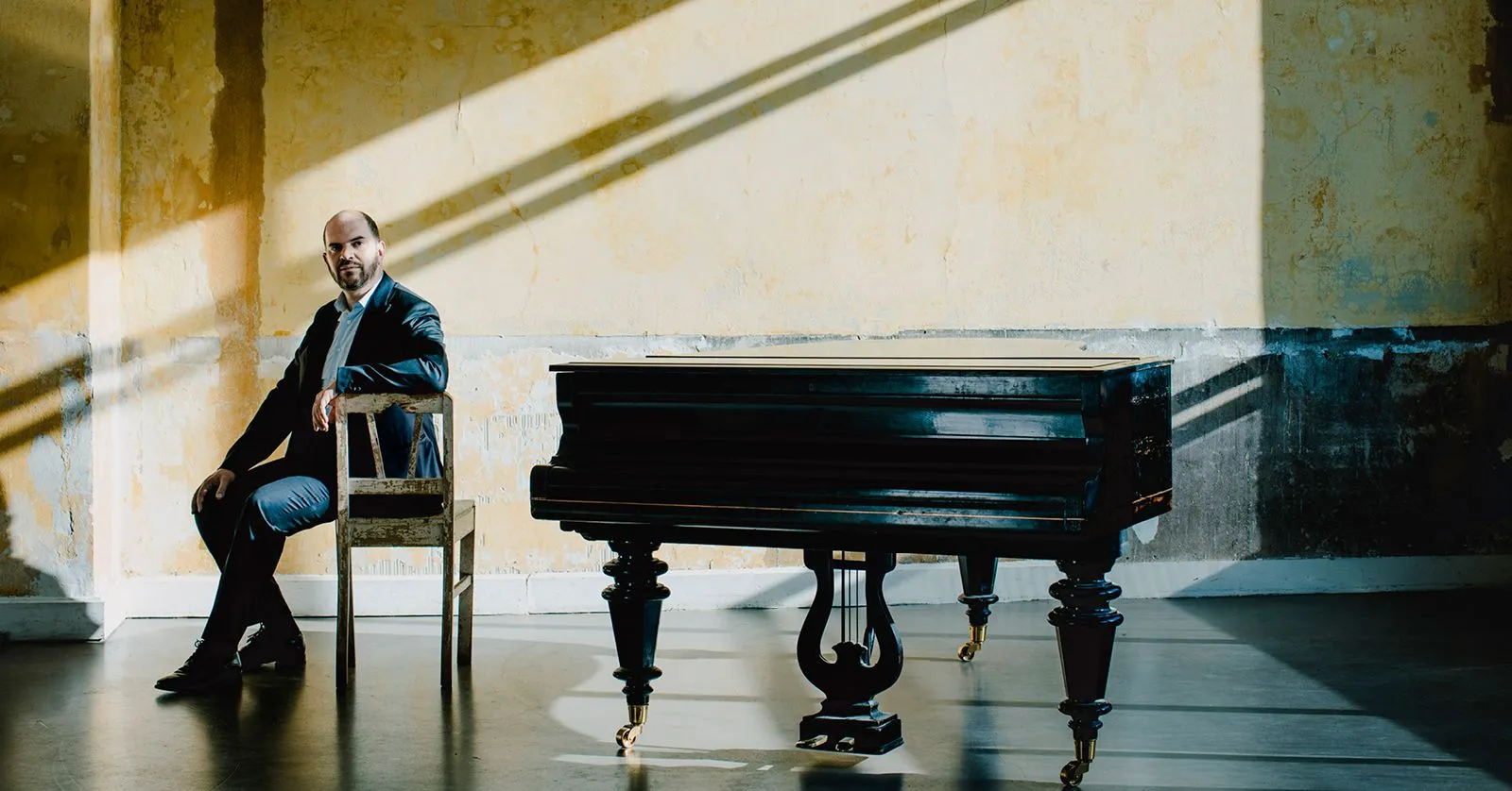 Kirill Gerstein é um homem branco, careca, de terno. Ele está sentado numa cadeira ao lado de um piano.