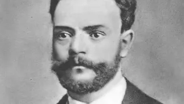 Retrato em preto e branco de Antonín Dvořák, com olhar intenso e barba cheia. Usa terno escuro com camisa branca, em frente a fundo neutro que destaca seu rosto e expressão séria.