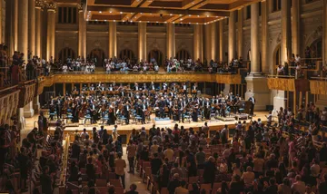 Os músicos da Orquestra estão na Sala de Concertos da Sala São Paulo. Todos vestem preto e estão segurando seus instrumentos.