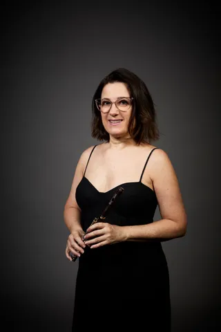 Fabíola Alves is a white woman with shoulder-length brown hair. She is wearing glasses, a black spaghetti strap dress, and holding a piccolo.