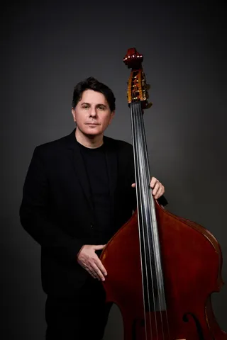 Jefferson Collacico is a white man with brown hair and a mustache. He is wearing a black T-shirt and holding a double bass.