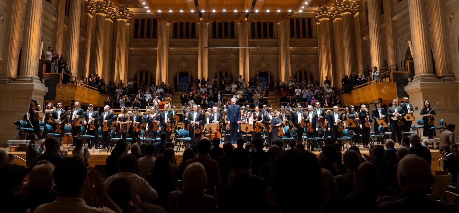 Foto da Osesp de pé no palco na Sala São Paulo com o maestro Thierry Fischer ao centro.